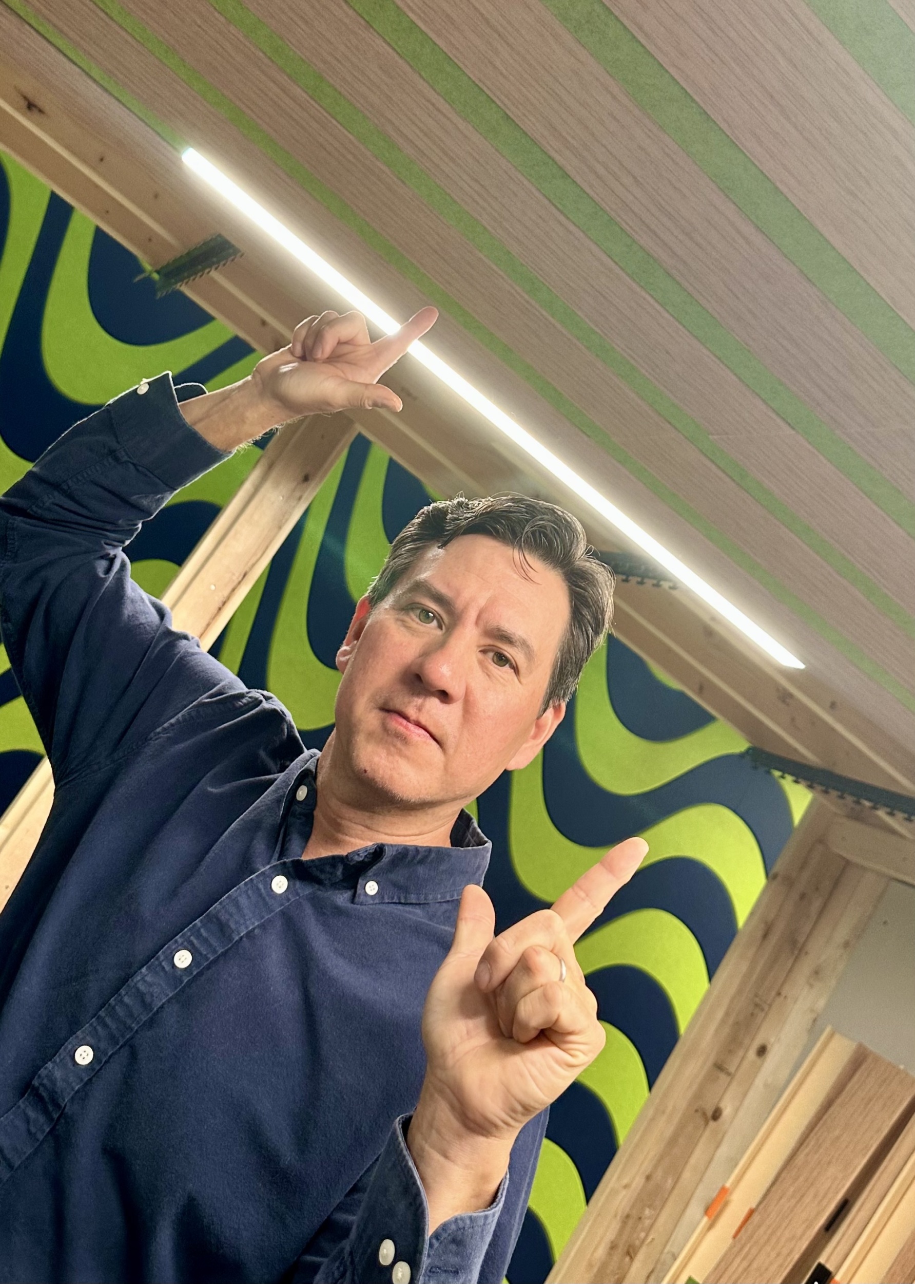 Man in a Blue Shirt Pointing Upward in a Room with Wooden Beams and Patterned Wall